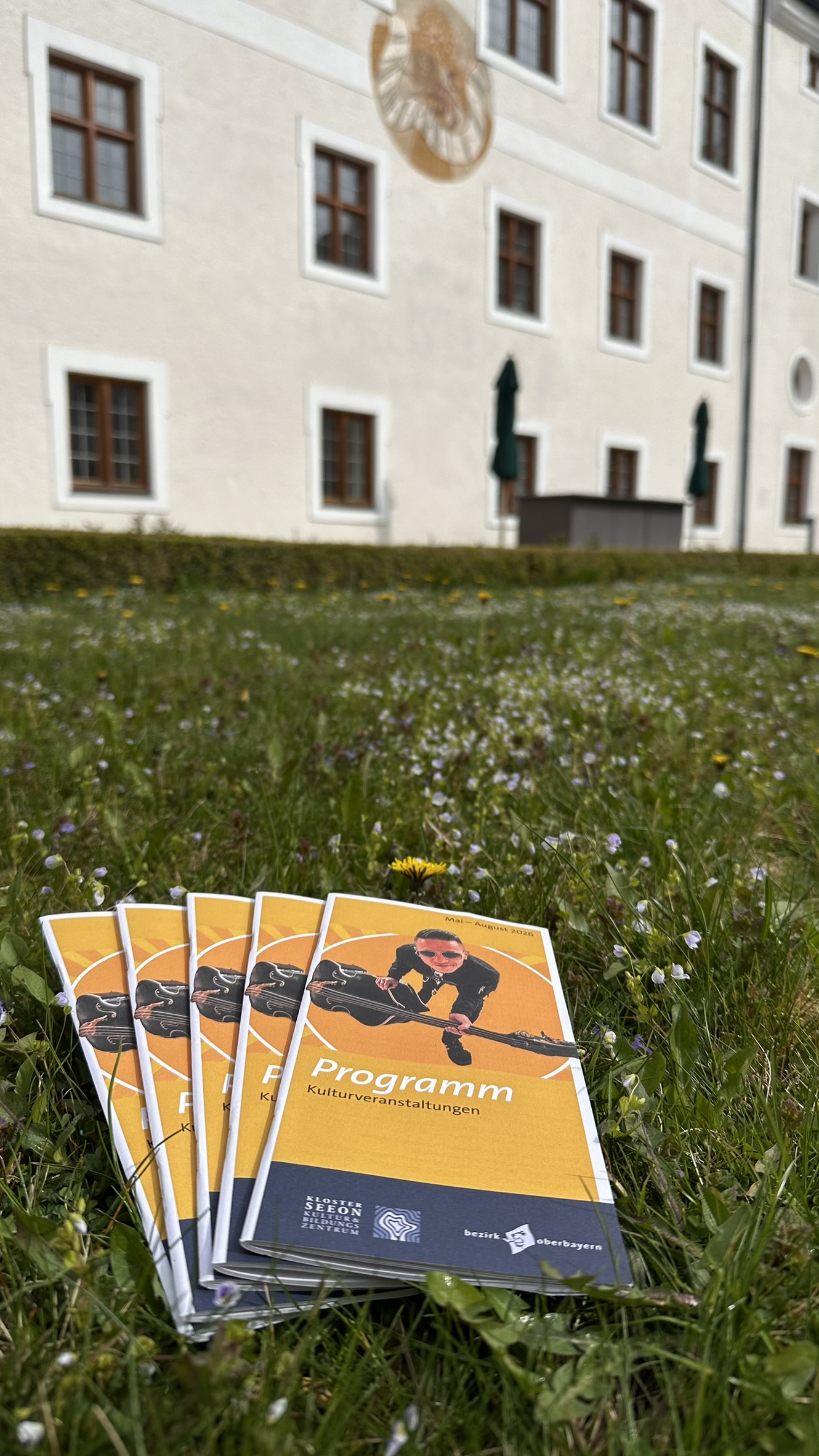 Programmhefte mit Illustration einer Person auf dem Cover liegen auf einer Wiese vor einem hellen Geb&auml;ude mit mehreren Fenstern und einer Sonnenuhr