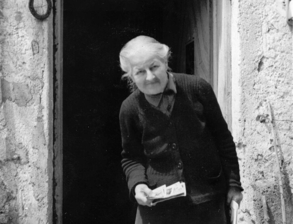 Die Schwarz-wei&szlig;-Fotografie zeigt eine alte, geb&uuml;ckte Frau, dunkel und eher &auml;rmlich gekleidet, vor einem alten Hauseingang.