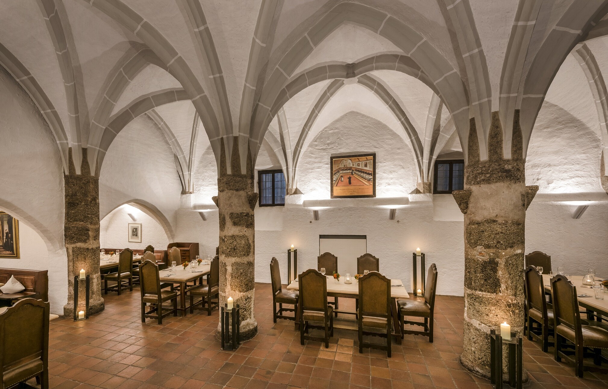 Ein Raum mit drei Steinsäulen in der Mitte, darüber ein gotisches Netzgewölbe, farblich hellgrau abgesetzt von den weißen Wänden; zwischen den Säulen Tische mit schweren Stühlen: Der Gotische Keller der Klostergaststätte Seeon.