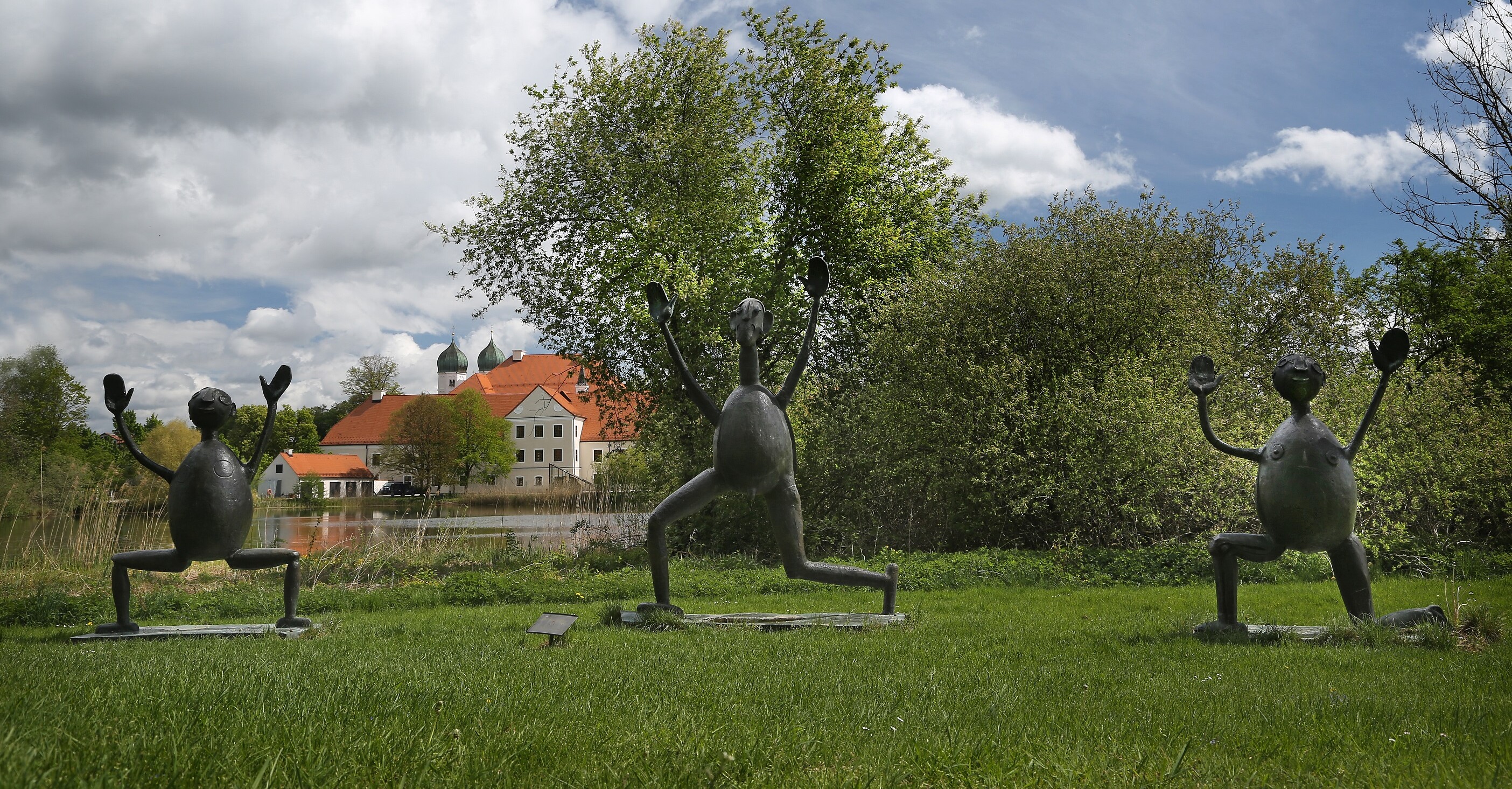 Drei überlebensgroße Bronzefiguren auf einer grünen Wiese, im Hintergrund Kloster Seeon