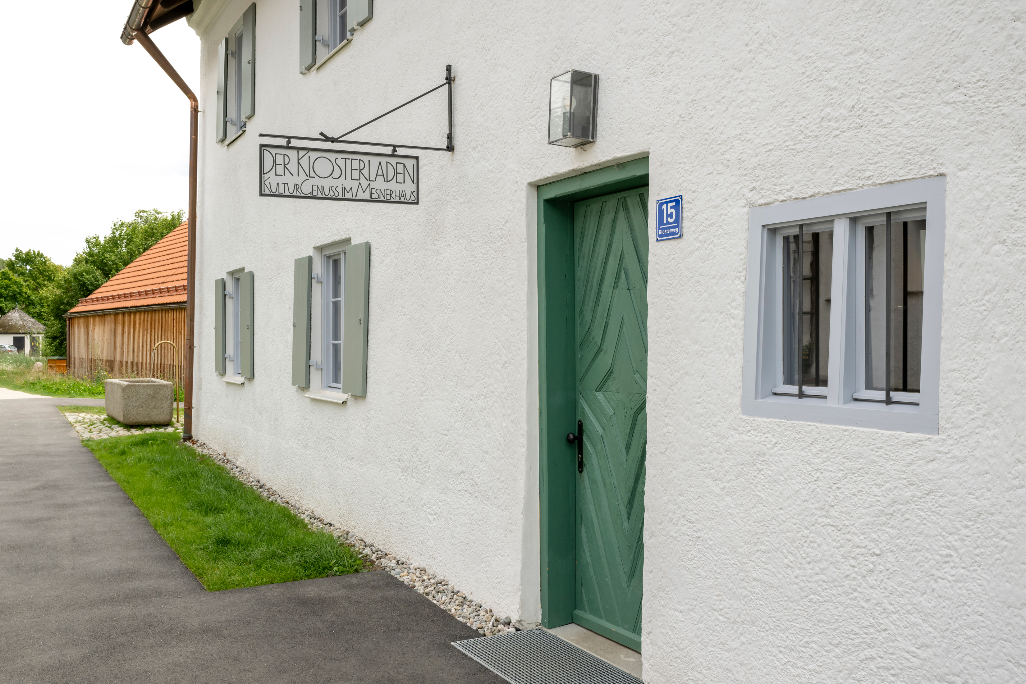 Ein Haus mit grüner Eingangstüre und Fenstern, an der Hauswand ist ein Schild angebracht worauf steht Der Klosterladen. KulturGenuss im Mesnerhaus