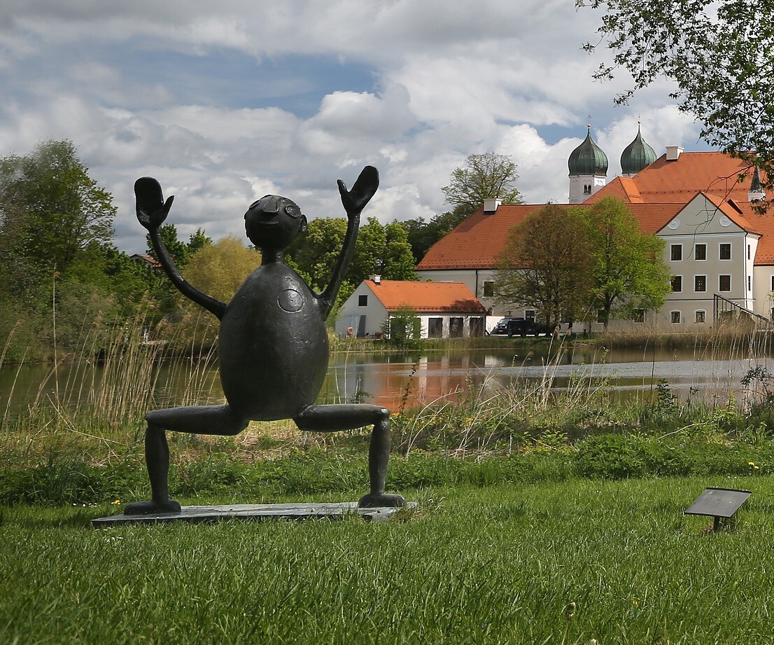 Eine Bronzefigur mit erhobenen Armen auf grüner Wiese, im Hintergrund Kloster Seeon