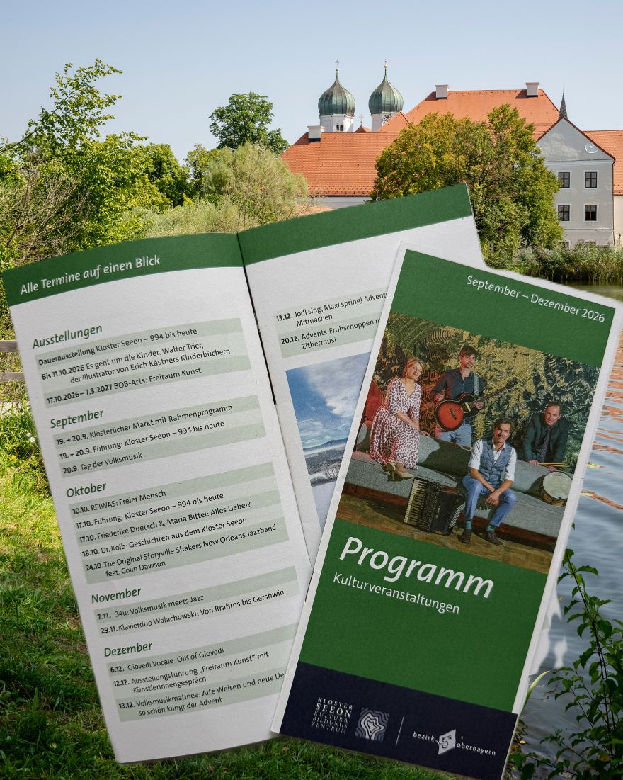 Aufgeschlagenes Veranstaltungsprogramm und Programmheft des Klosters Seeon f&uuml;r September bis Dezember 2026 vor einer Au&szlig;enaufnahme des Klosterensembles am See. Im Hintergrund sind die Geb&auml;ude des Klosters mit roten Ziegeld&auml;chern, zwei markanten Zwiebelt&uuml;rmen sowie B&auml;ume und Wasser zu sehen. Auf dem aufgeklappten Programm sind Termine f&uuml;r Ausstellungen, Konzerte und weitere Kulturveranstaltungen aufgef&uuml;hrt.
