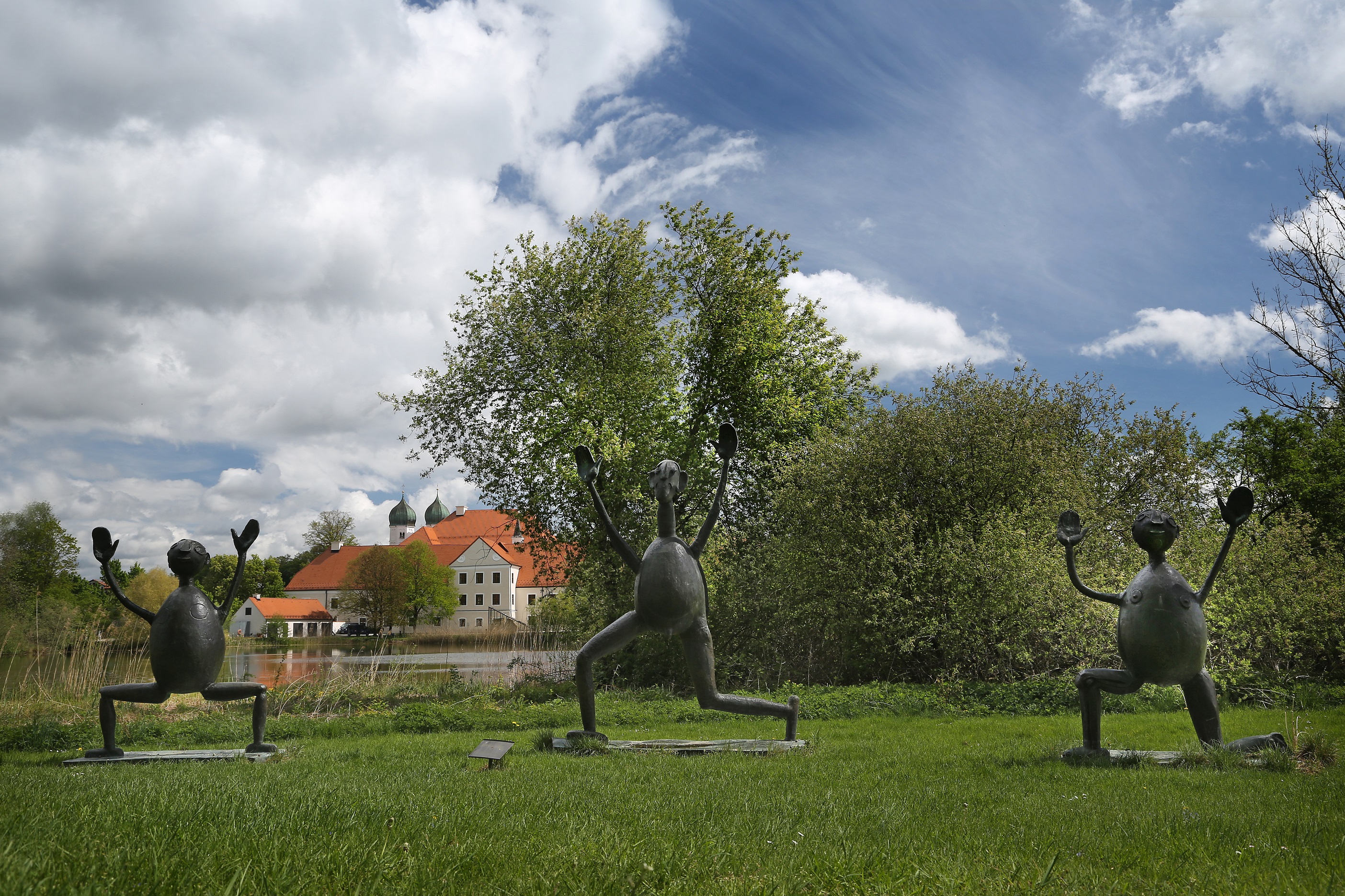 Drei Bronzefiguren mit erhobenen Händen auf einer Wiese, im Hintergrund Kloster Seeon