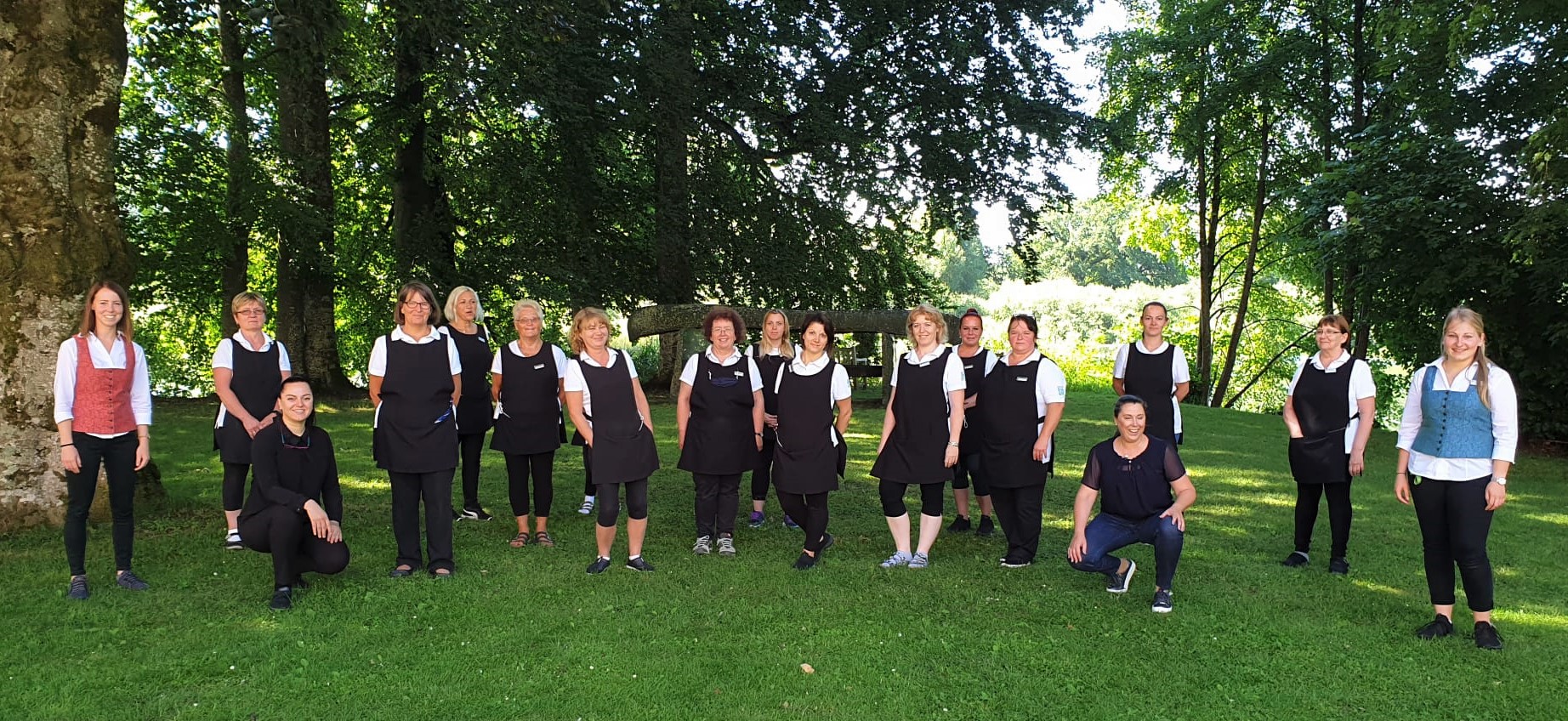 Das Team vom Housekeeping in Kloster Seeon: Eine Gruppe von Frauen steht auf einer grünen Wiese. Im Hintergrund sind Bäume zu sehen .Die Frauen haben weiße T-Shirts und schwarze Schürzen. Links eine Dame mit rotem Mieder, rechts eine Dame mit blauem Mieder. 