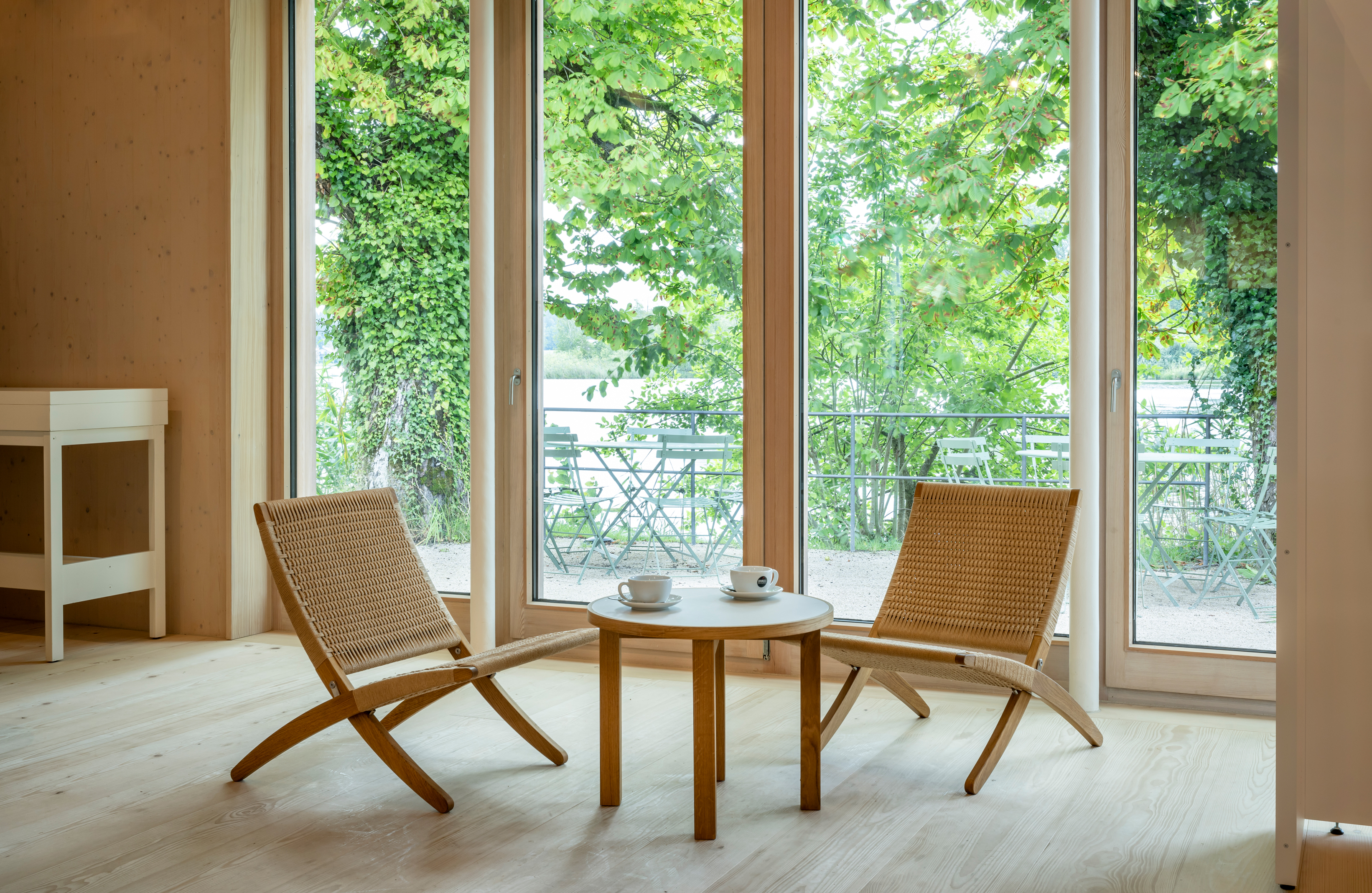 Sitzgruppe aus hellem Holz und Korb mit zwei Stühlen und einem kleinen Tisch vor bodentiefen großen Fenstern, draußen grünes Laub