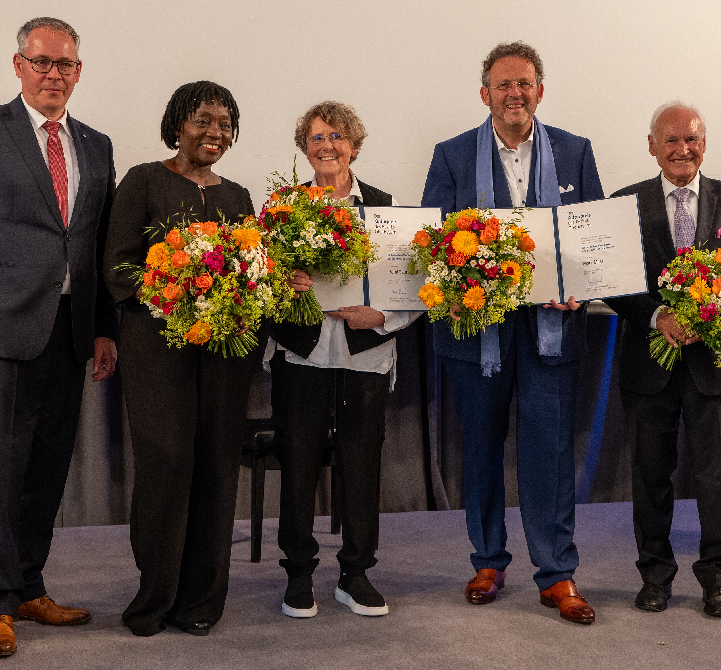 Oberbayerischer Kulturpreis 2024: Bezirkstagspräsident Thomas Schwarzenberger  mit den Laudatoren und Preisträgern, die jeweils Blumen und Urkunden in den Händen halten.