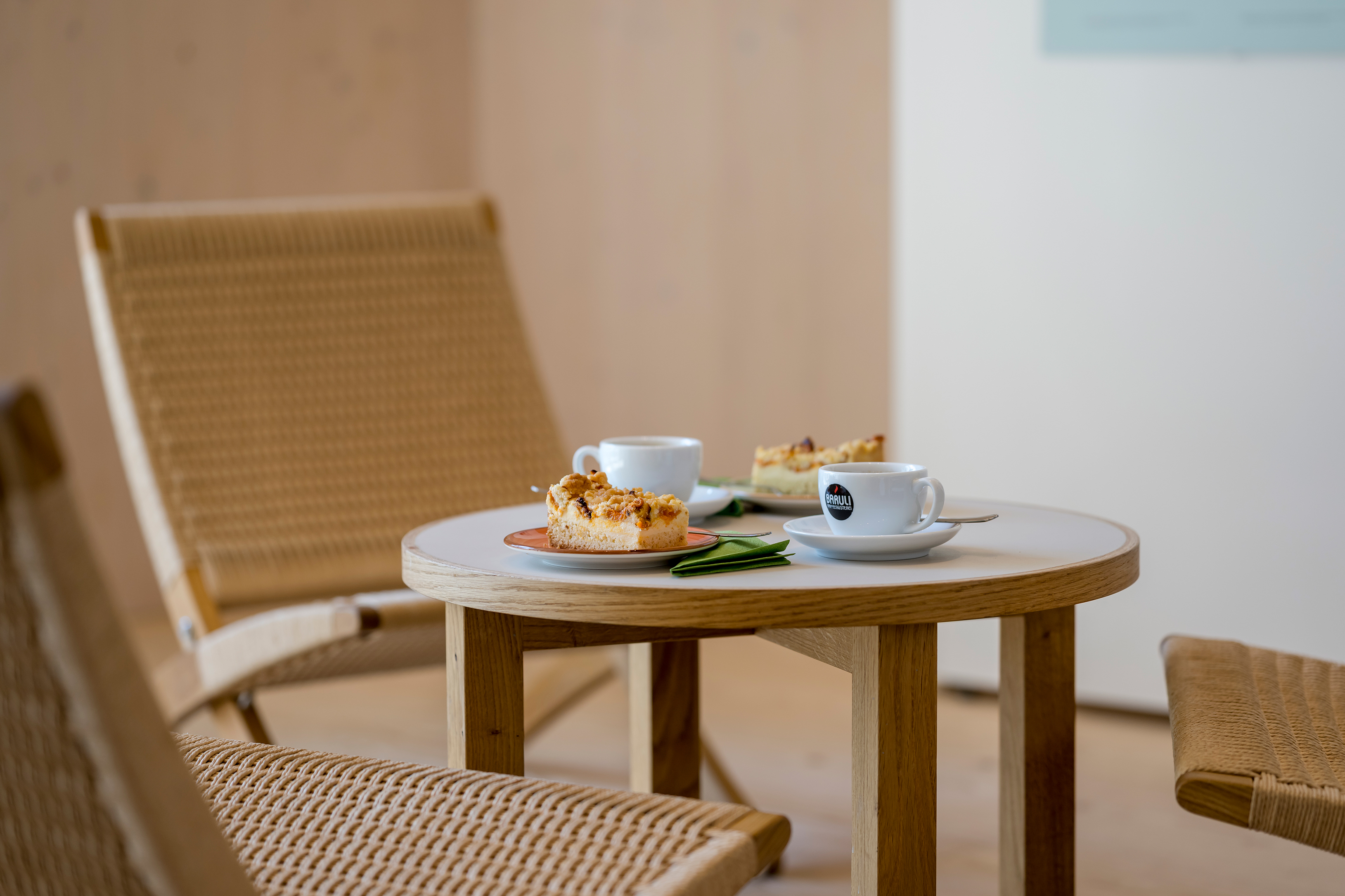 ... bei einer gemütlichen Tasse Kaffee oder Tee im Ausstellungsbereich ... Sitzgruppe aus hellem Holz und Korb mit zwei Stühlen und einem kleinen Tisch vor bodentiefen großen Fenstern, draußen grünes Laub