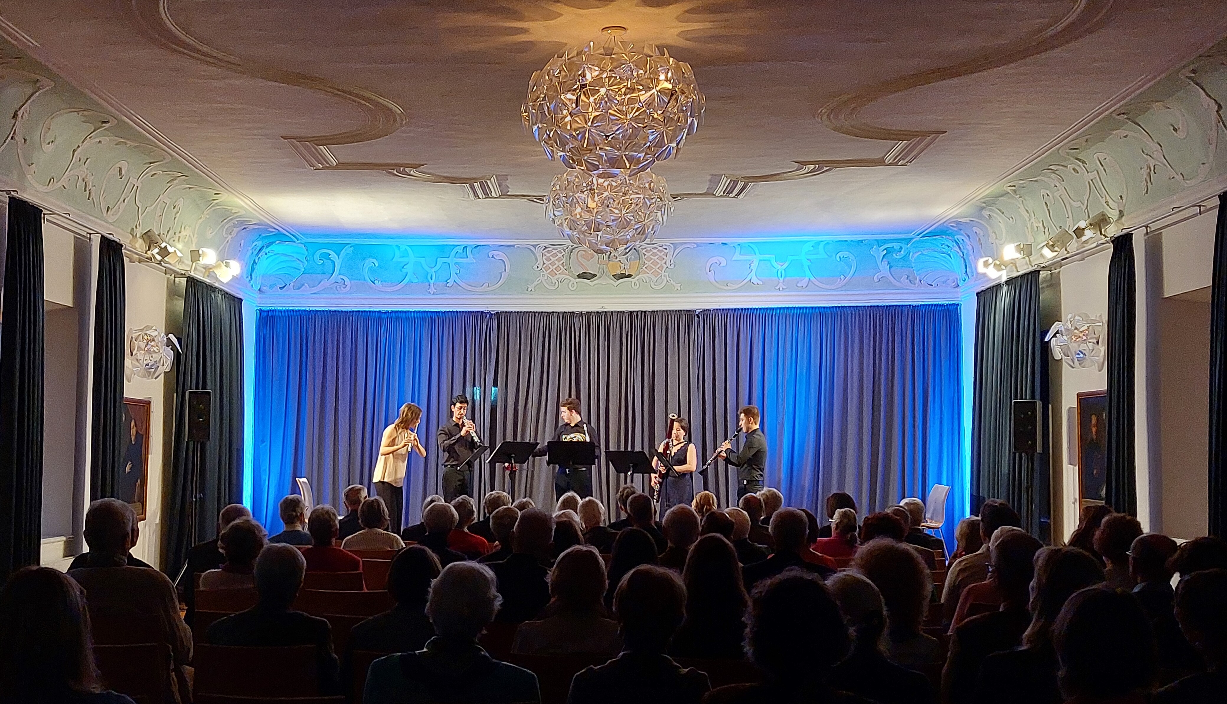 Blick aus dem dunklen Publikumsraum auf die hell erleuchtete Bühne im Festsaal mit vier jungen Musikern in der Mitte