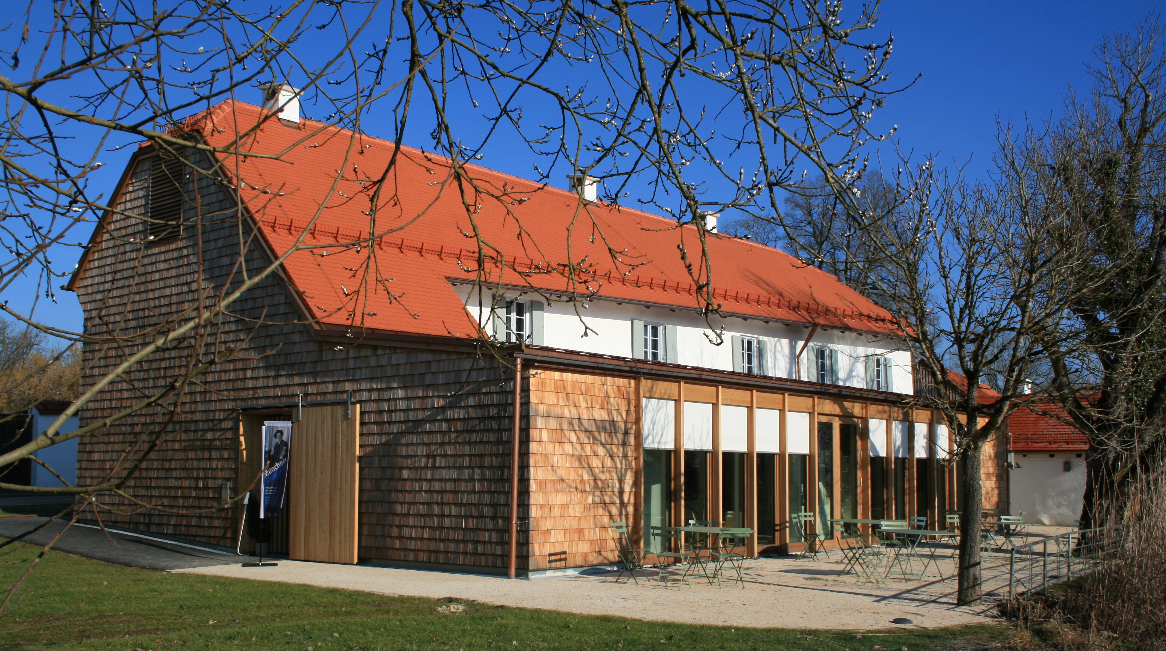 Das denkmalgeschützte Mesnerhaus bei Kloster Seeon, im Vordergrund Zweige mit Knospen, dahinter die Terrasse des Hauses mit grüner Bestuhlung