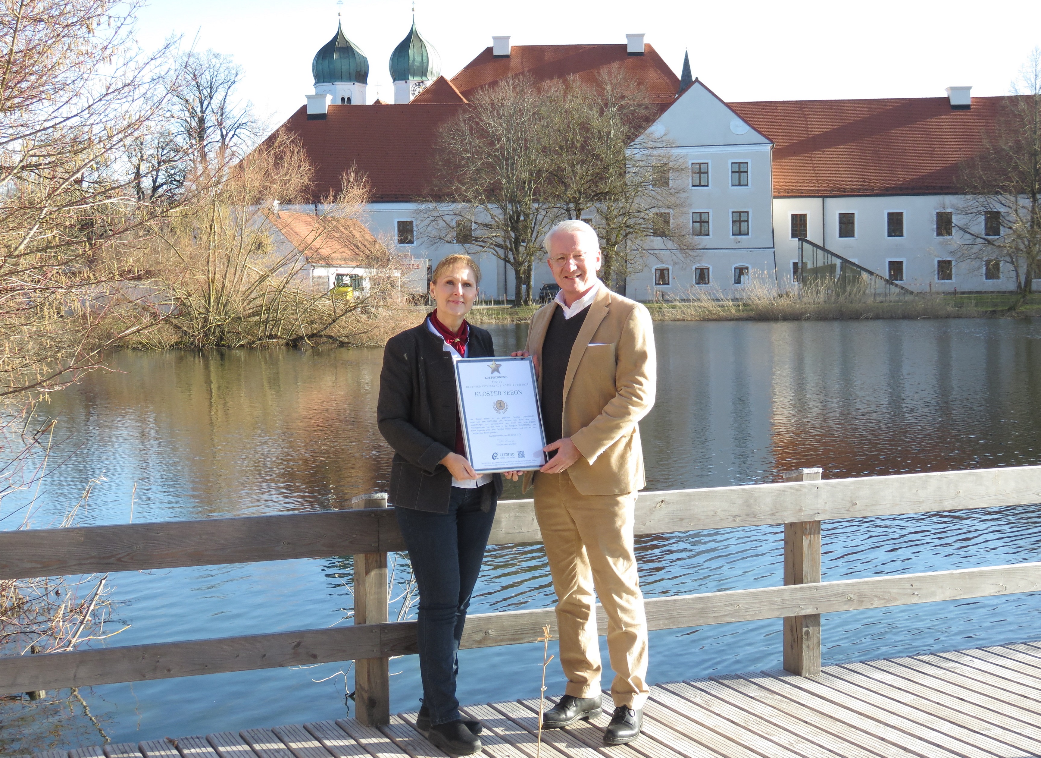 Inge Ederer-Posch und Gerald Schölzel (Geschäftsleitung Kloster Seeon) stehen auf einem Steg vor dem Fachzentrum Fisch & Biene und halten beide die Urkunde in den Hängen, im Hintergrund sind die beiden Zwiebeltürme von Kloster Seeon und die ehemalige Klosteranlage zu sehen.