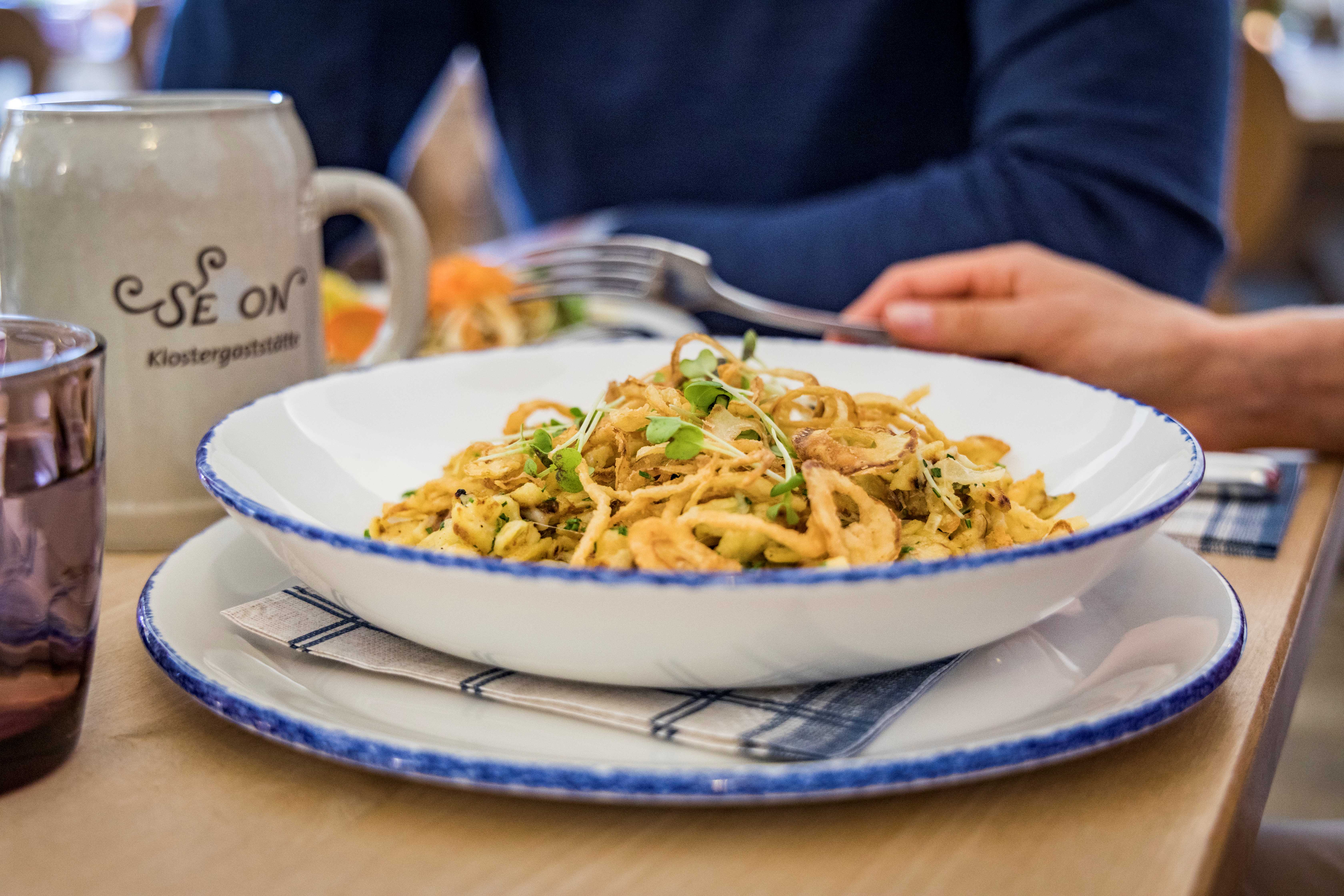 ein deftiges Gericht, angerichtet auf einem blau weißen Teller, daneben ein Kloster-Seeon-Bierkrug auf einem Holztisch in der Klostergaststätte Seeon