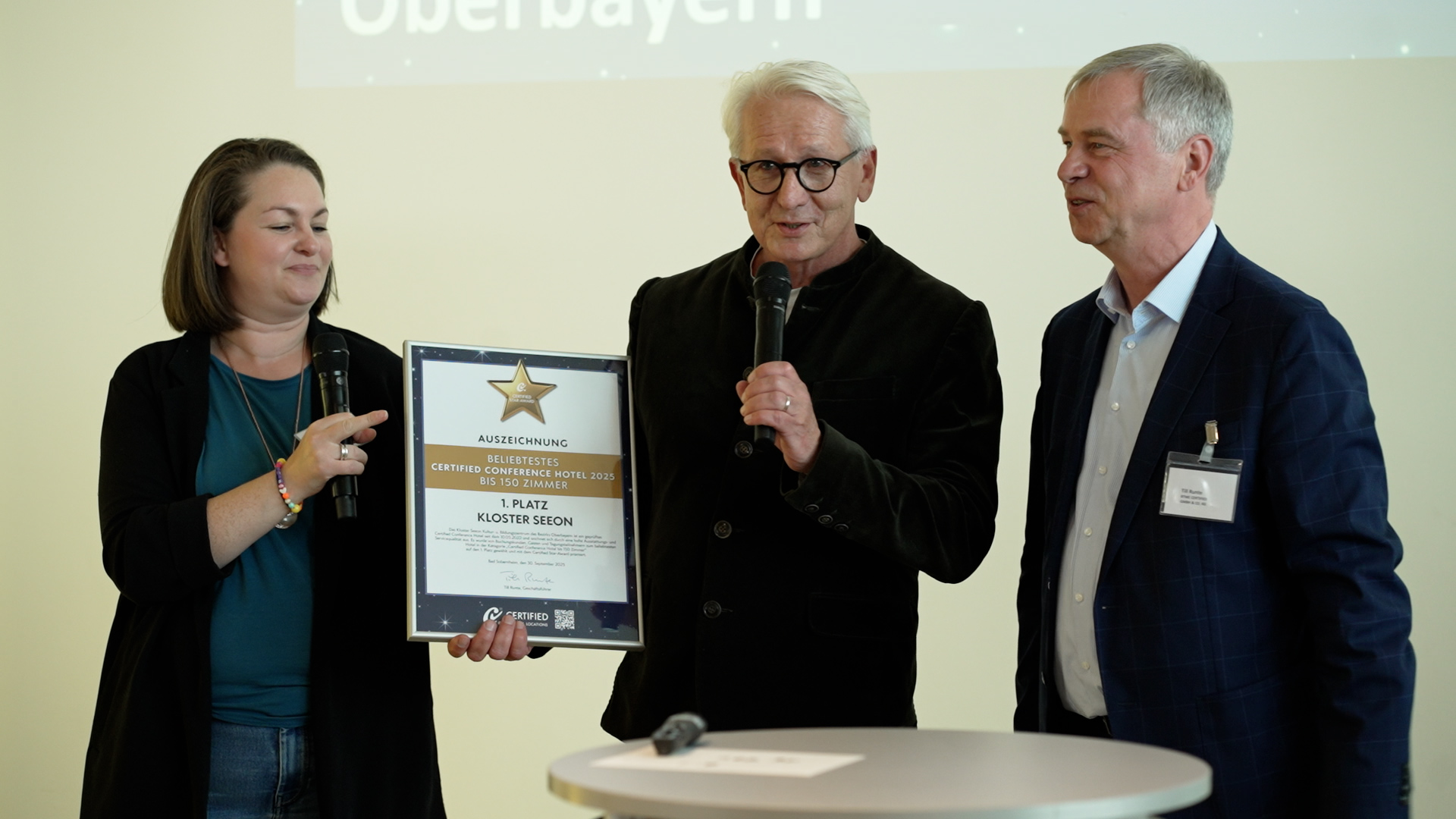 Drei Personen Inga Beerenrath, Gerald Schölzel, Till Runte bei der Preisverleihung. Herr Schölzel hält ein Mikrofon und die Urkunde in der Hand.