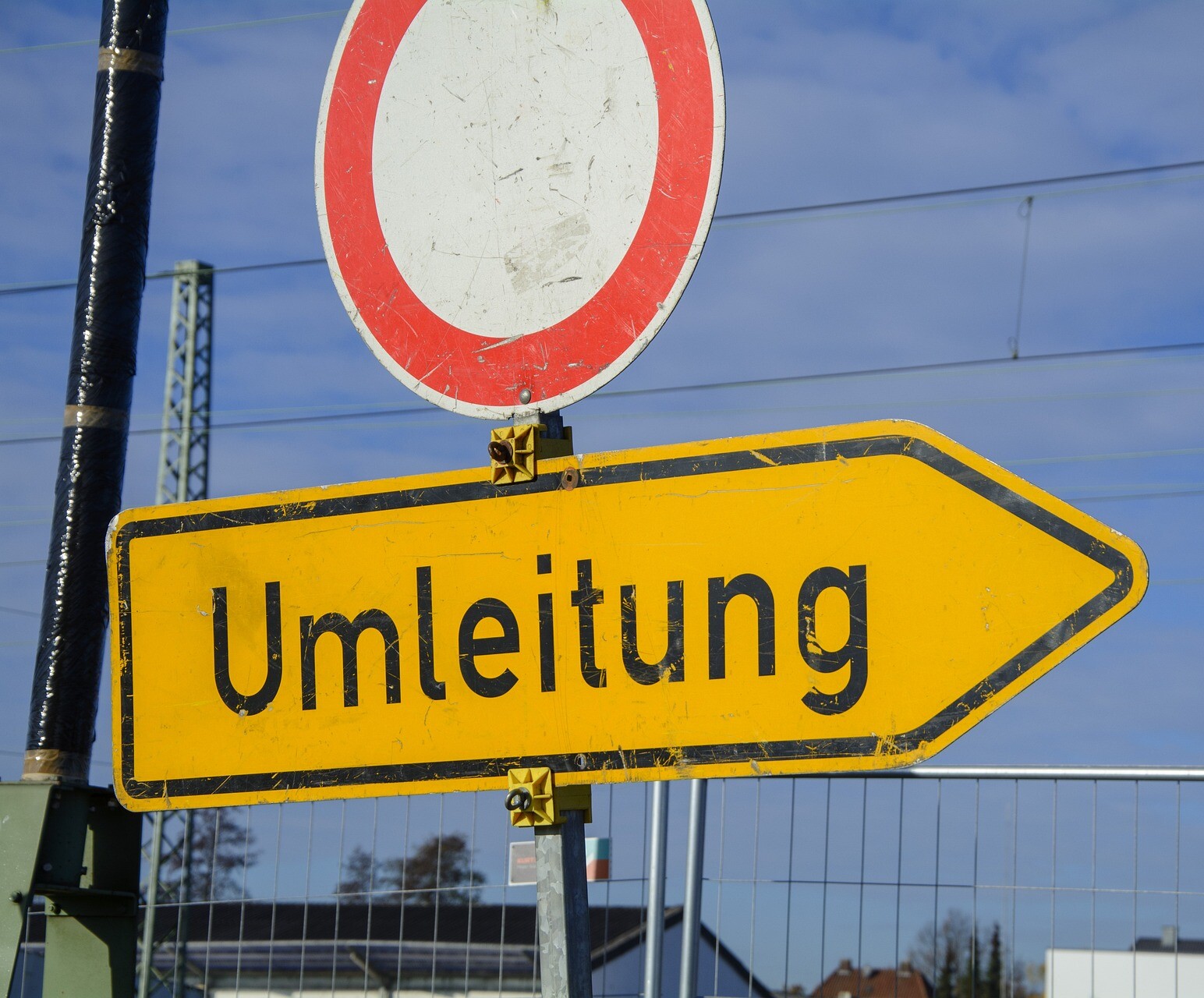 Ein gelbes Schild in Pfeilform mit der schwarzen Aufrisft Umleitung, im Hintergrund der obere Teil eines Bauazauns, darüber ein rundes Schild mit roter Umrandung