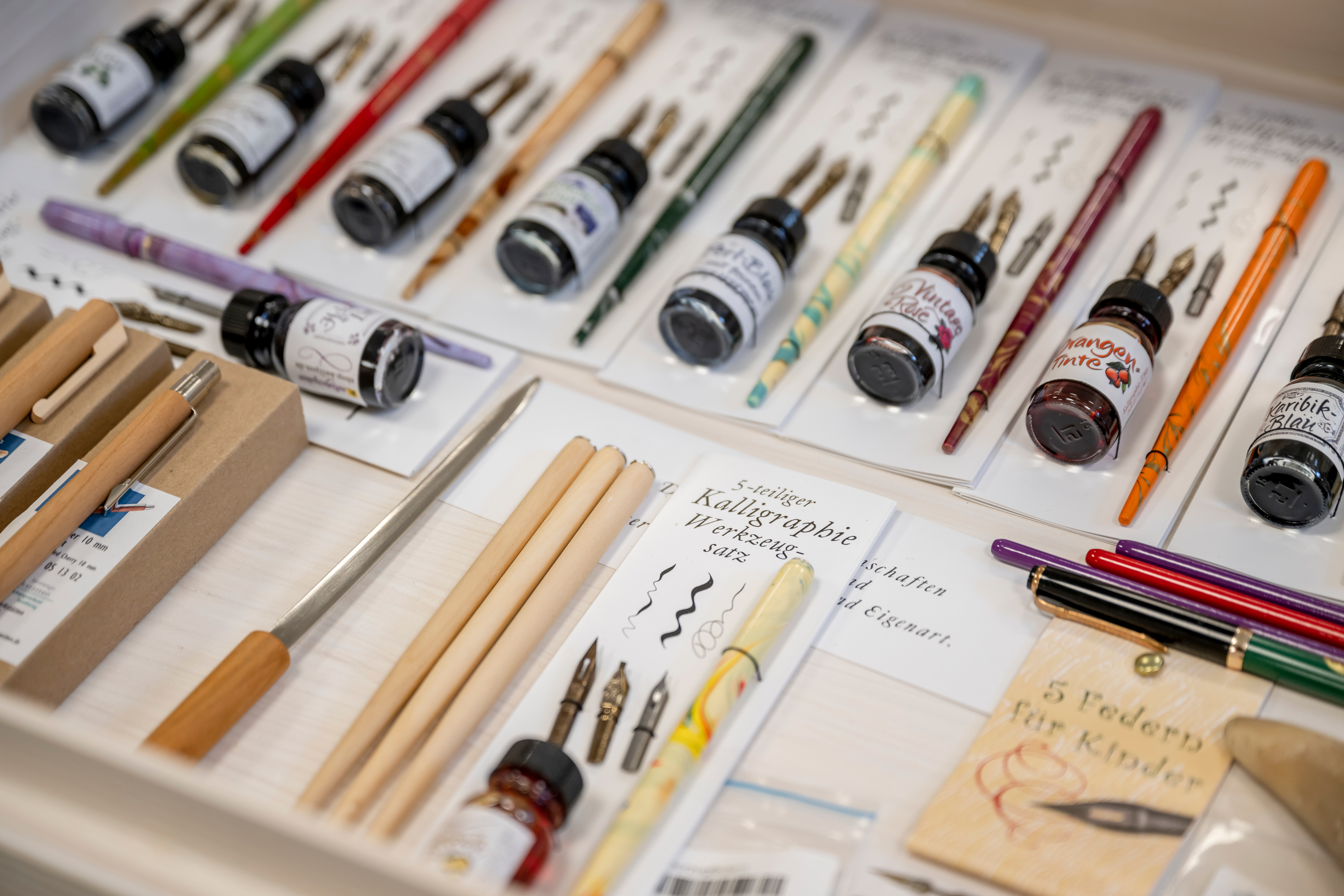 Geöffnete Schublade aus hellem Holz, darin verschiedene bunte Federkiele und Schreibfedern, kleine Gläser mit verschiedenfarbiger Tinte und Stifte