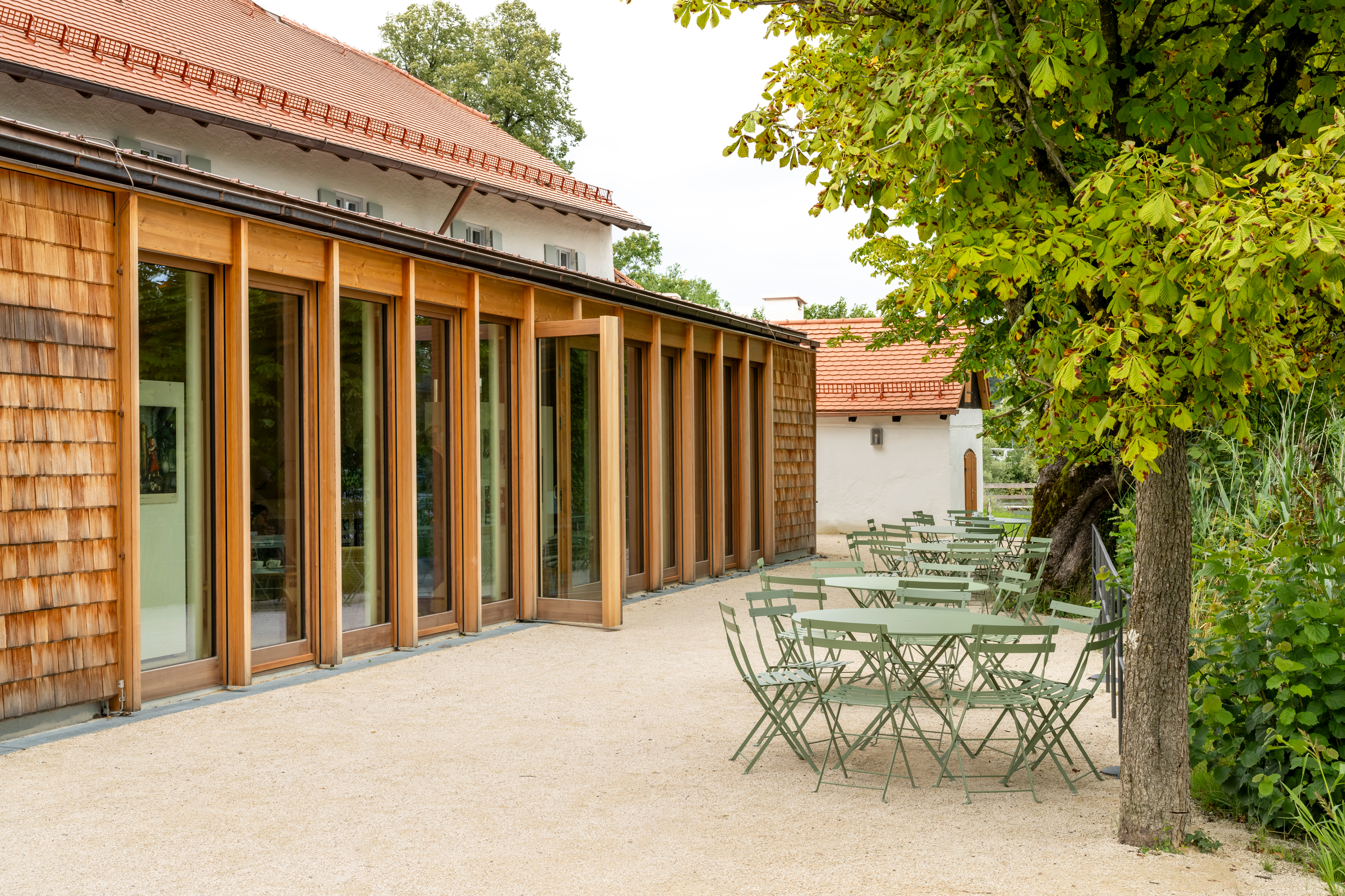 Blick auf die Terrasse des Mesnerhauses, rechts vorne runde Gartentische und Stühle in grüner Farbe, links der Wintergarten des Mesnerhauses aus Holz mit geöffneter Tür zur Terrasse hin